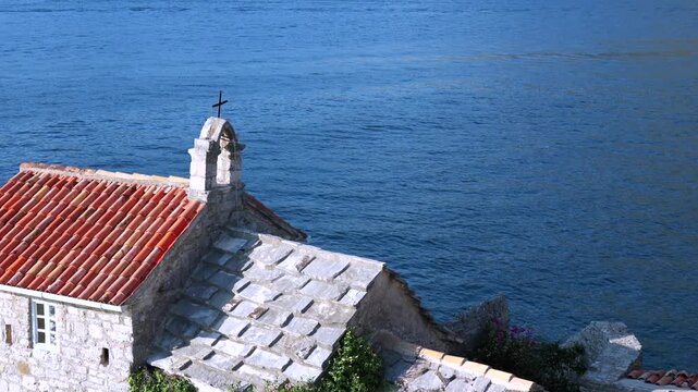 Old Stone Church by the Sea. Old stone chapel roof with a small cross and red clay tiles overlooking blue sea on a sunny Mediterranean coast.