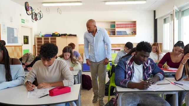 High school teacher walking through the classroom and monitoring a group of multiracial teenage students taking an important exam, ensuring a fair and controlled testing environment