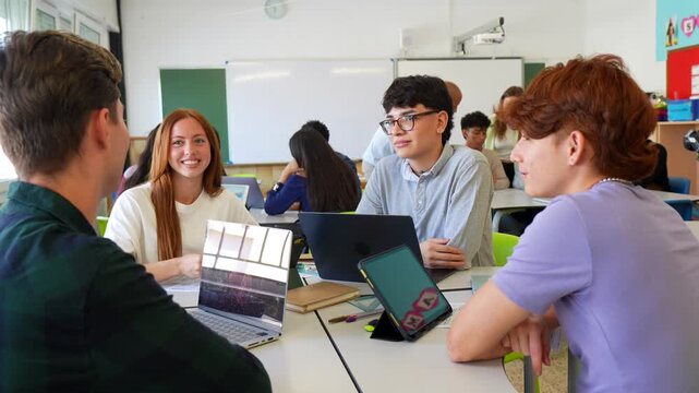 Diverse group of teenage students talking and working together on a collaborative assignment in a modern classroom, using laptops and tablets to research and share ideas for their school project