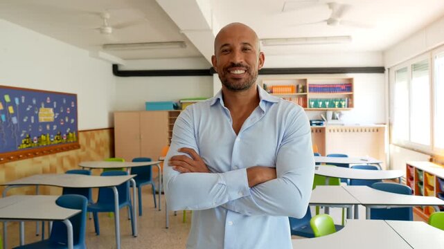 Handsome male professor with a friendly smile standing with arms crossed in an empty classroom, ready for the beginning of the school year and looking confidently at the camera
