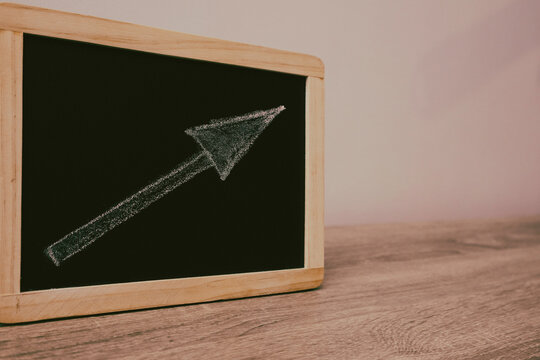 Chalkboard with white chalk arrow pointing up right on wooden desk surface representing growth development and business success