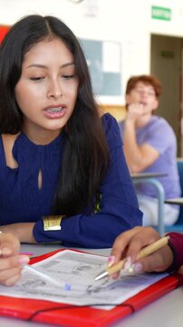 Young female student with braces discussing and working on a group project with a classmate at her desk, concentrating on the shared document during an educational lesson in a high school classroom
