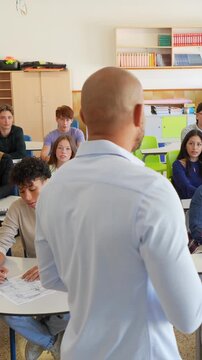 Group of attentive multiethnic teenage students listening to their male professor during a class, focusing on education and learning while sitting at their desks inside a modern classroom