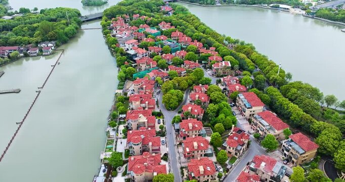 Aerial view of residential villas with red roofs surrounded by green trees along a river under bright daytime lighting.