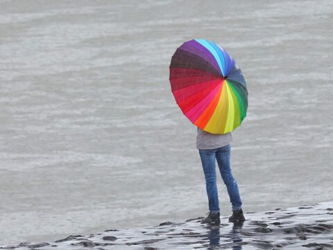 Vue de derri&egrave;re d'une femme seule devant la mer  sombre et portant un parapluie multicolore dans une ambiance pluvieuse. Symbole LGBT et de solitude.
