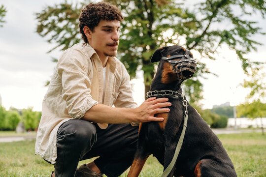 Young man kneeling beside a Rottweiler dog wearing a muzzle in a grassy park setting with trees and a cloudy sky in the background