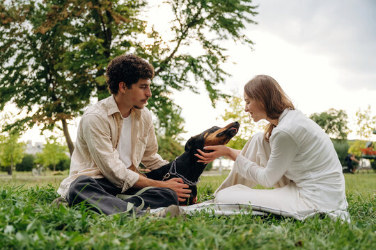 Young couple sitting on grass in a park, interacting with a black dog, surrounded by trees and a cloudy sky in the background