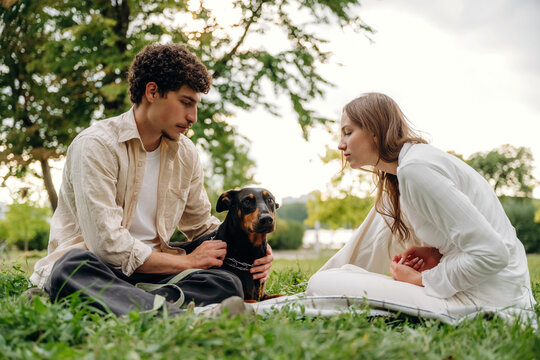 Young couple sitting on grass in a park, interacting with a black dog, surrounded by trees and greenery under a cloudy sky