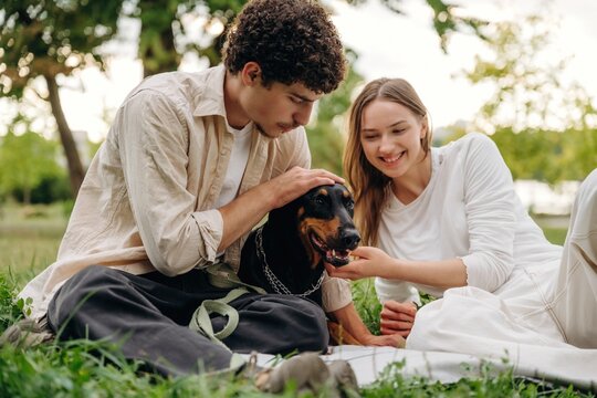 Young couple sitting on grass in a park, interacting with a black dog, surrounded by trees and greenery on a sunny day