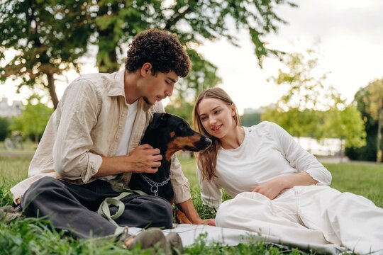 Young couple sitting on grass in a park, interacting with a black and tan dog, surrounded by trees and greenery on a sunny day