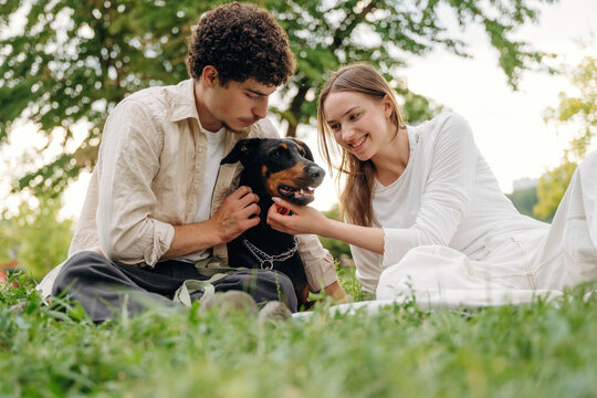 Young couple sitting on grass in a park, interacting with a black dog, surrounded by trees and greenery, enjoying a sunny day outdoors