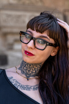 Alternative woman posing, showing neck and chest tattoos, lip piercing, and glasses