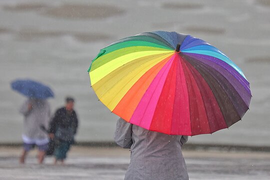 Vue rapproch&eacute;e d'un parapluie multicolore abritant une personne seule sous un temps pluvieux avec deux personnages floues en arri&egrave;re plan