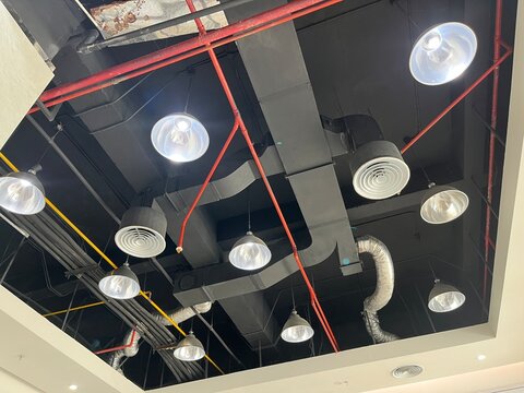 Modern industrial ceiling featuring black air ducts red pipes and hanging metal pendant lamps.