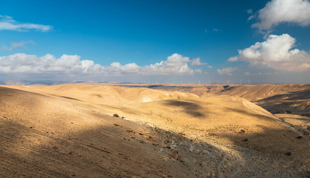 View of the arid desert landscape near Shobak Castle with distant wind turbines under a blue sky with scattered clouds in Shobak, Ma'an Governorate, Jordan.