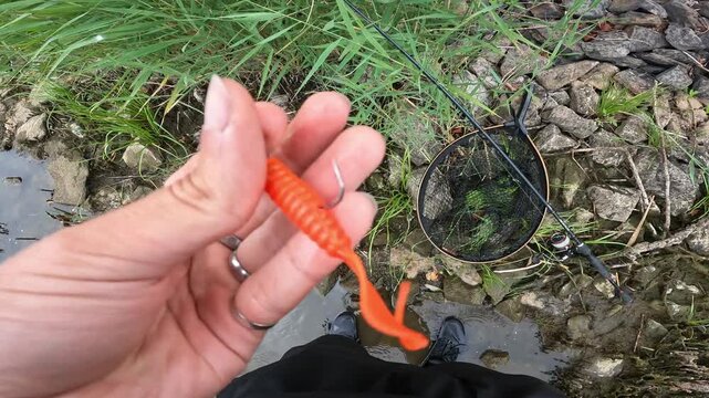 Close-Up Male Hand Holding Orange Soft Plastic Lure on Jig Head. Fishing Tackle and Net on River Bank.