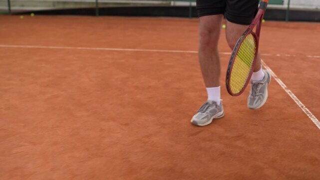 Caucasian young adult man walking across clay court holding tennis racket in slow motion with bouncing ball. Ideal for sports apparel advertising, tennis club promos, athletic training campaigns