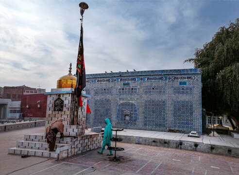 View of the Tomb of Shah Yusuf Gardezi with its intricate blue tiled walls and golden dome, featuring a person in a green shawl walking in the courtyard in Multan, Punjab, Pakistan.