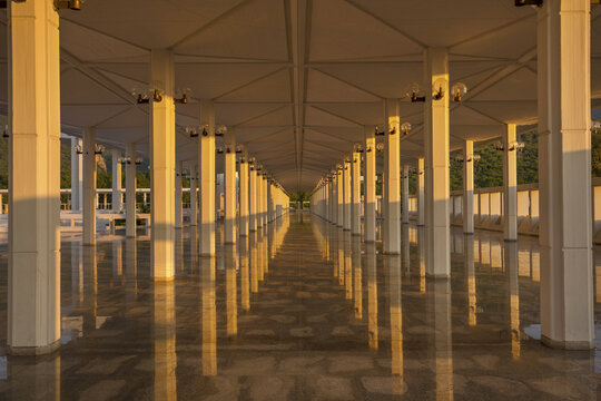 View of the symmetrical colonnade at Faisal Masjid with white pillars and reflective stone floors illuminated by warm golden hour light in Islamabad, Islamabad Capital Ter, Pakistan.
