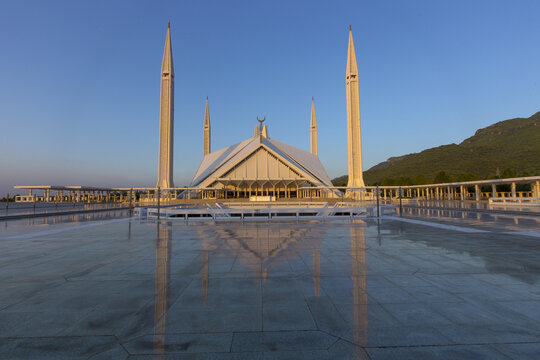 View of Faisal Masjid with its distinctive tent-like structure and four tall minarets reflecting on the polished courtyard floor at sunset in Islamabad, Islamabad Capital Ter, Pakistan.