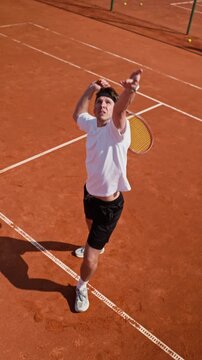Vertical video of Caucasian young adult man bouncing ball and performing jump serve on clay tennis court in slow motion. Footage suited for sports advertising and fitness campaigns