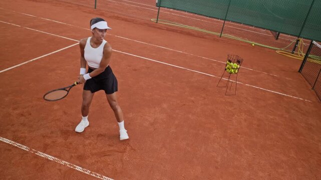 Biracial young adult woman dropping ball and hitting two handed backhand on clay court in slow motion from high angle. Suitable for sports marketing and tennis training campaigns
