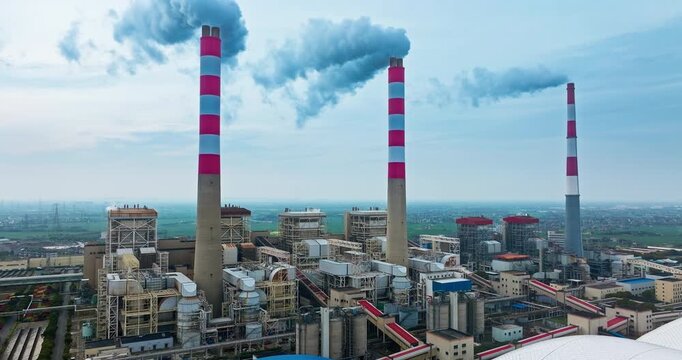Aerial view of a thermal power plant with smoke emitting from tall striped chimneys under bright daylight.