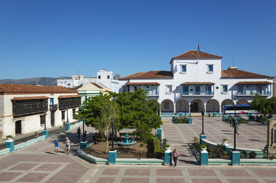 View of Parque Cespedes with the historic Ayuntamiento building, trees, a fountain, and people walking in the square under a clear blue sky in Santiago de Cuba, Santiago de Cuba, Cuba.
