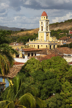 View of the iconic yellow bell tower of the Convento de San Francisco de Asis rising above the colonial rooftops and lush green trees in Trinidad, Sancti Spiritus, Cuba.