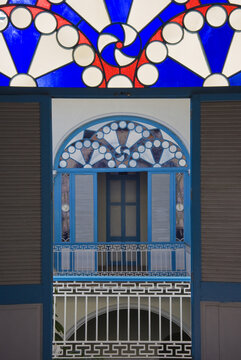 View of a colonial-style interior balcony featuring vibrant blue and red stained glass fanlights, wooden shutters, and intricate white metal railings in Havana, Havana, Cuba.