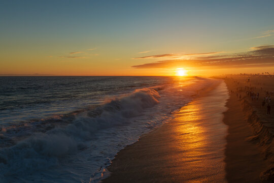 Golden sunset over Newport Beach, California, with powerful Pacific Ocean waves rolling toward the sandy shoreline as warm sunlight reflects across the wet beach and silhouettes beachgoers along the c