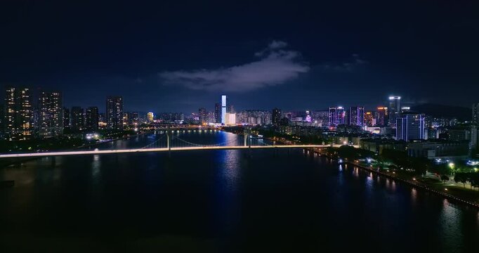 Aerial view of illuminated cityscape at night, featuring a bridge spanning across a wide river and modern skyscrapers under artificial lighting.