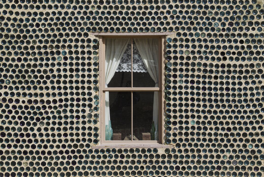 View of the historic Bottle House wall constructed from glass bottles surrounding a window with white lace curtains in Rhyolite, Nevada, United States.
