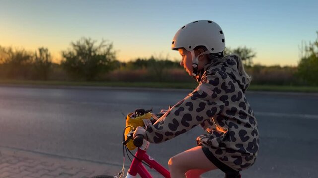 Young cyclist pedals pink bike beside empty road in golden evening light. White helmet, hooded jacket, and steady posture show safe travel outdoors as rider moves past low shrubs and open sky.