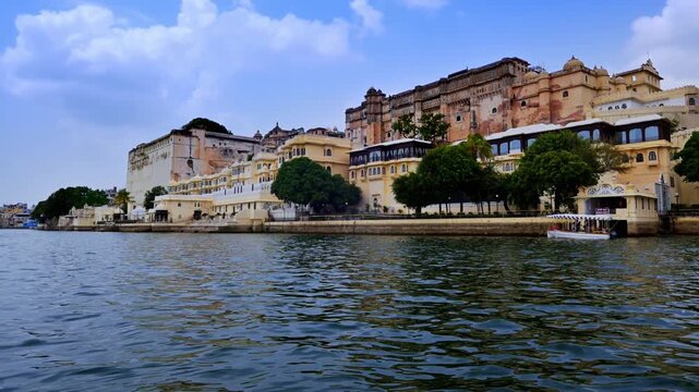 A moving waterfront view reveals pale and ochre buildings, balconies and shaded terraces along the City Palace complex in Udaipur.