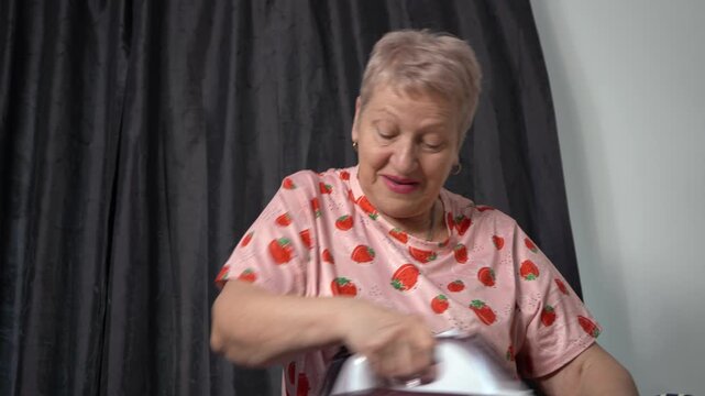 Elderly woman ironing clothes with an iron on a board. Grandmother chores routine. Tell and talk