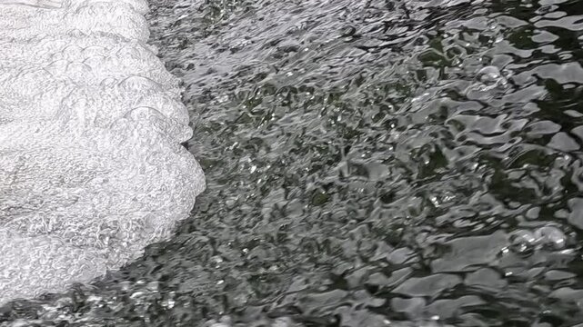 Close-up view of white foam and bubbles washing steadily over a dark wet rocky surface, moving across the frame as water flows and swirls gently over the stones near the shore.