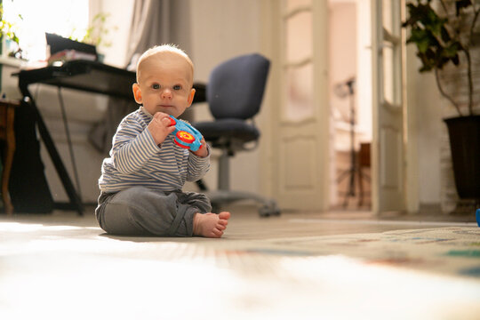 Cute baby in a striped shirt sitting on a rug, playing with a colorful toy in a bright, sunlit room