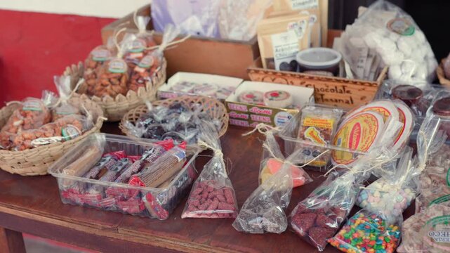 Close-up panning shot of artisanal Mexican sweets and colorful candies at a Tapalpa market stall, featuring rustic packaging, vivid treats, and inviting ambient daylight.