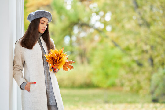 Autumn portrait of a beautiful happy smiling woman with yellow maple leaves in a gray beret in the park