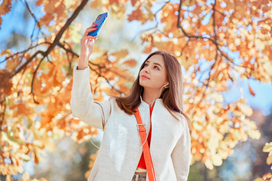 A young beautiful woman walks through the autumn park. A girl with a smartphone takes photos in an autumn forest