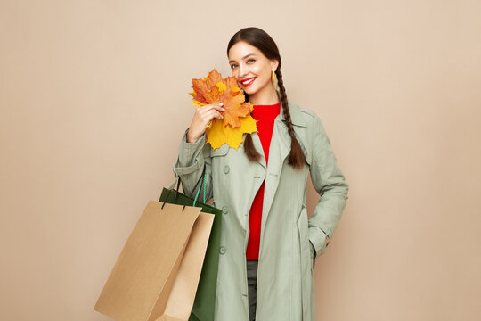 Autumn portrait of beautiful happy young woman with shopping bag, stylish girl holding yellow maple leaves in round hat, coat on beige studio background