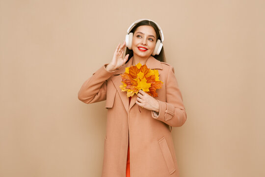 A beautiful young woman in a fashionable autumn outfit listens to music with wireless headphones. A girl in a stylish orange blouse poses on a beige background