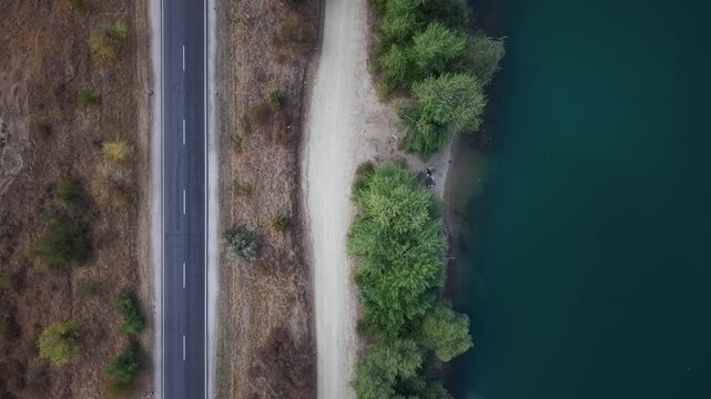 An overhead aerial view shows an empty asphalt highway running parallel to a lush shoreline and deep turquoise lake water.