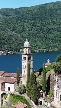 Morcote Switzerland vertical drone orbit around lakeside church bell tower, cypress trees and turquoise Lake Lugano, Ticino alps.