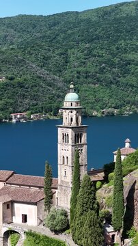 Morcote Switzerland vertical drone shot of lakeside church bell tower framed by cypress trees, turquoise Lake Lugano below.