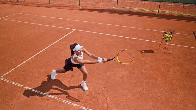 Young adult woman hitting tennis balls with two handed backhand on clay court in high angle. Perfect for sports marketing, athletic training, and fitness campaigns