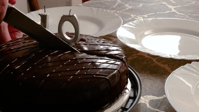 A horizontal video of a large knife cutting into a glazed chocolate birthday cake next to white ceramic plates on a table.