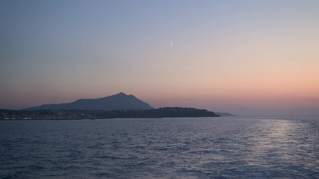 Calm evening light glows over the sea and Procida&rsquo;s distant coastline as a thin crescent moon hangs in the pastel sky.