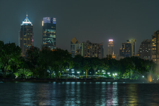 View of the illuminated skyline of Bangkok rising above the lush trees and calm waters of Benjakitti Park at night in Krung Thep Maha Nakho, Thailand.